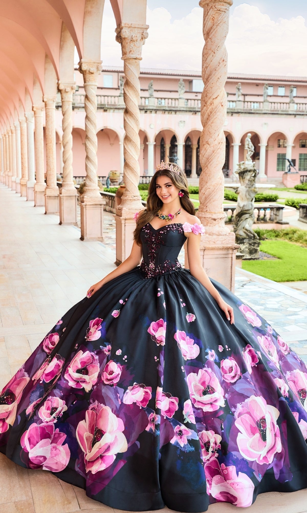 A woman in a tiara wears the Pink Floral Black Quinceanera Dress: Princesa PR30151, featuring bold pink floral designs, as she stands in an elegant outdoor corridor with columns and a manicured garden.